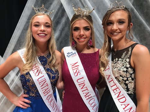 Three girls wearing tiaras, one in blue, one in red, one in black, all wearing sashes that read "Miss America Miss Uintah County 2018"