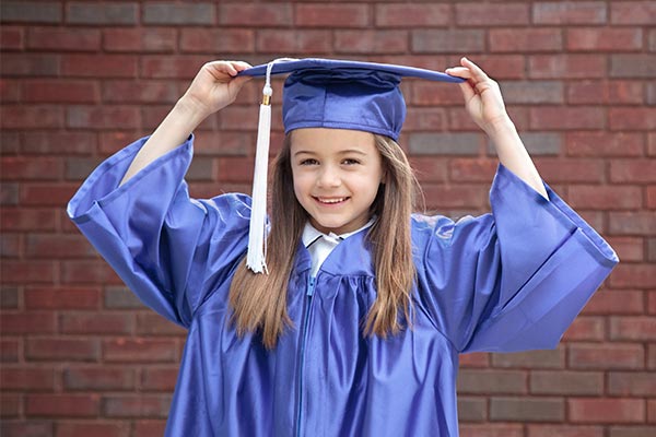 Little girl with brown hair wearing blue graduation gown and cap holding the brim of cap and standing in front of a brick wall.