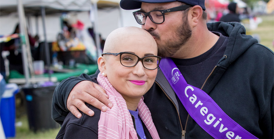 Man with black glasses, hat, and jacket wearing a purple "Caregiver" sash and kissing the bald head of a woman who survived cancer, wearing black glasses and pink scarf