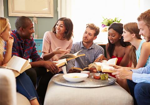 seven men and women sitting in a living room talking with books