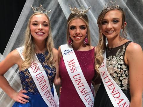 Three girls wearing tiaras, one in blue, one in red, one in black, all wearing sashes that read "Miss America Miss Uintah County 2018"